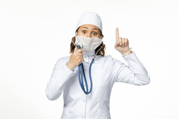 正面正面图年轻女医生戴防护无菌口罩因冠状病毒使用听诊器在白色表面专业灭菌医疗
