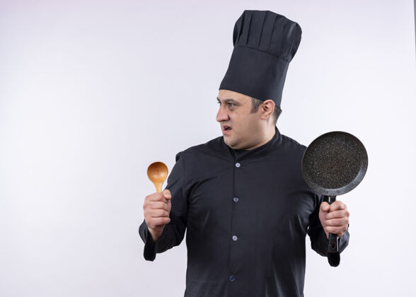 厨师男厨师身穿黑色制服 头戴厨师帽 手拿木勺和平底锅 站在白色背景下惊讶地向一旁望去站着黑木头