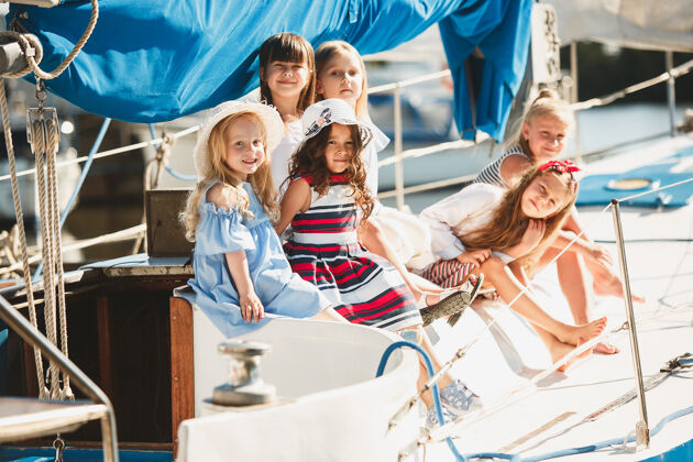 姐妹孩子们在游艇上喝着橙汁青少年或少女们在蓝天下户外五颜六色的衣服孩子们的时尚 阳光明媚的夏天 河流和假日的概念年轻人水手衣服