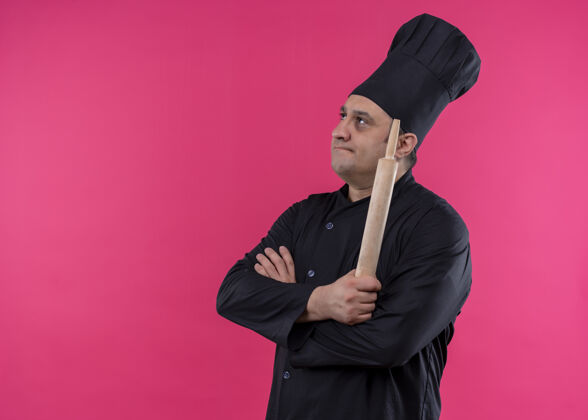 思想体贴的男厨师身穿黑色制服 头戴厨师帽 手持擀面杖 面带沉思的表情站在粉色背景上站着烹饪粉色