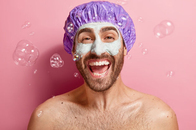 裸体室内拍摄表情满意的男人用泥塑面膜 享受淋浴和面部护理 戴浴帽 肥皂泡四处飞舞 洗身大笑乐观卫生