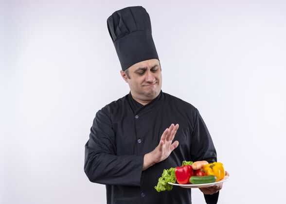 站立男厨师身穿黑色制服 头戴厨师帽 手持盛有新鲜蔬菜的盘子 站在白色背景上做着带着怀疑表情的防御手势怀疑白色黑色