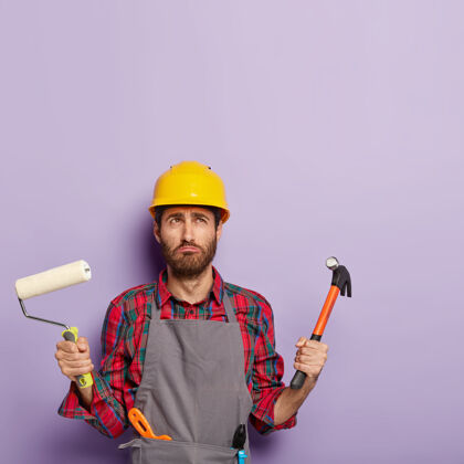 模型不满的男子戴着防护安全帽 围裙 手持油漆辊和锤子 忙于房屋装修 手持劳保工具紫色翻新年轻