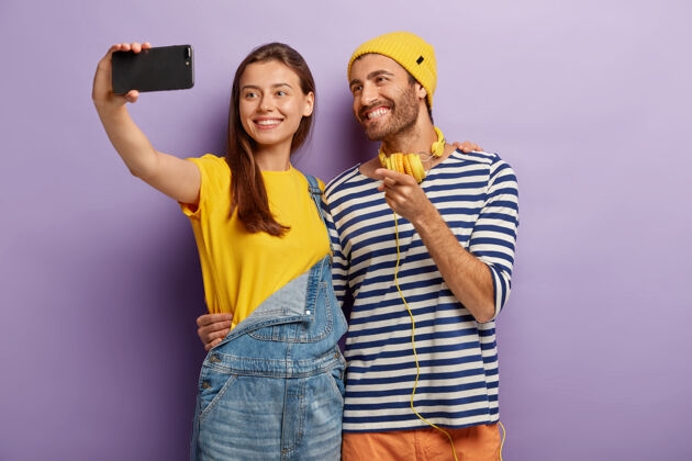 情人快乐的男女青少年在智能手机上自拍 微笑着拥抱 拥抱着对方 穿着时髦的衣服 站在紫色的墙上 指着展台 自拍使享受手机