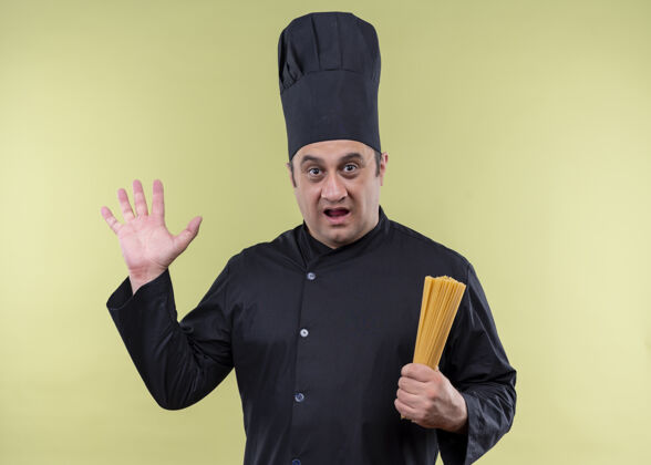 厨师男厨师身穿黑色制服 头戴厨师帽 手拿一排意大利面 举手站在绿色背景上 显得既惊讶又惊讶看黑色男