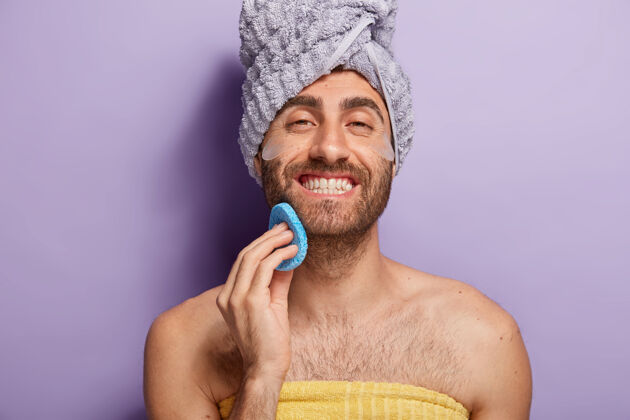 化妆品快乐的年轻人洗澡后整理自己 想要有干净的皮肤 拿着美容海绵贴在脸上 裹着毛巾 面带微笑湿巾男性紫色