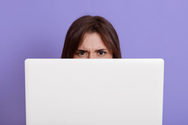 休闲装模特躲在笔记本电脑后面 隔着淡紫色的墙 脸上有愤怒的表情 黑头发的女人躲在白色的笔记本后面 工作时是自由职业者成人网络空间服装