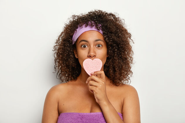 赤裸上身照片中的年轻美籍黑人女子戴着紫色的头巾和毛巾围在身上 用化妆心形海绵捂住嘴 擦脸非洲化妆品年轻化