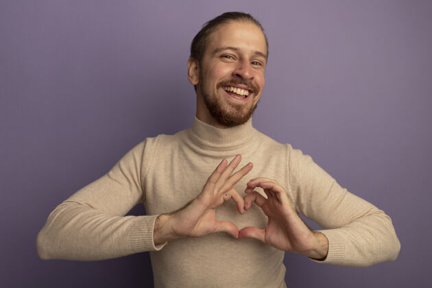 男人穿着米色套头衫的年轻帅哥看着前面 高兴地微笑着 用手指在淡紫色的墙上做着心形手势表情淡紫色帅气