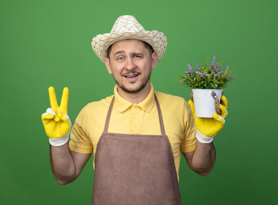 市民年轻的园丁穿着连体衣 戴着工作手套 手里拿着盆栽植物站在绿色的墙上绿色工人连身衣