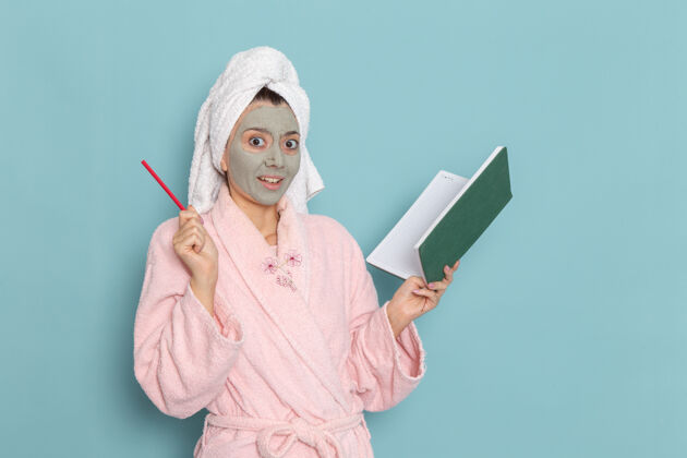 特纳正面图身穿粉色浴袍 脸上戴着口罩的年轻女性拿着蓝色墙壁上的抄写本淋浴清洁美容自护霜抄写本专业正面