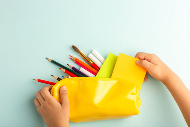 小的俯视图：在冰蓝色的表面上 一个小孩拿着装满彩色铅笔的黄色笔盒钢笔画画书写工具