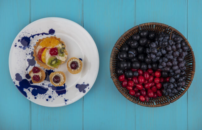 甜食在盘子里的水果蛋糕和水果的顶视图 如山茱萸浆果和葡萄在蓝色背景上盘子树莓葡萄