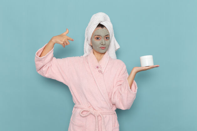 举行正面图年轻女性穿着粉色浴袍淋浴后脸上戴着口罩拿着面霜在蓝色墙壁上淋浴清洗美容自理霜观看人自我照顾