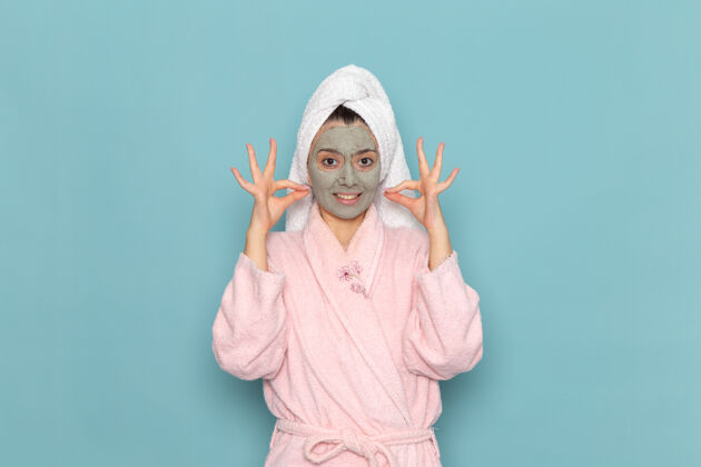 面具正面图身着粉色浴袍的年轻女性 脸上戴着口罩 在蓝色墙上清洗美容自护霜淋浴视图男人清洁