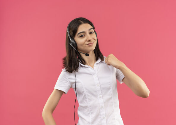 女孩穿着白衬衫戴着耳机的年轻女孩 面带微笑地看着前方握紧拳头 站在粉红色的墙上微笑麦克风手势