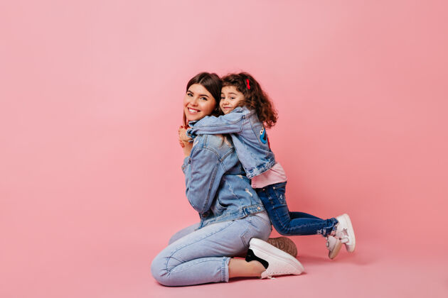 幼儿园可爱的孩子拥抱在粉红色背景上的母亲幸福的妈妈和小女儿在牛仔裤工作室拍摄黑发拥抱童年