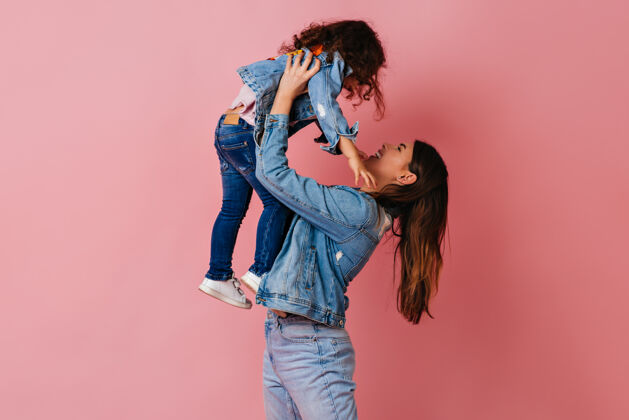 幸福浅褐色的年轻女子抱着女儿 背景是粉红色的摄影棚拍摄的妈妈和穿着牛仔夹克的未成年孩子女孩妈妈幼儿园