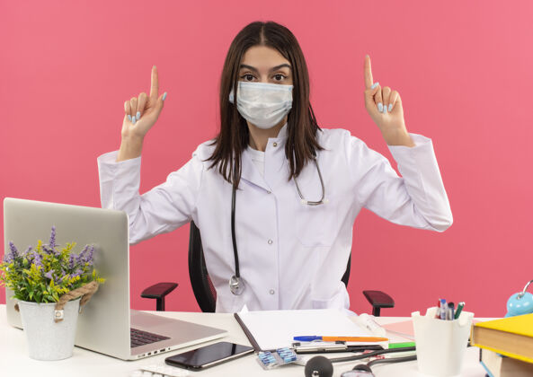 脖子年轻的女医生穿着白大褂 戴着面罩 脖子上戴着听诊器 食指朝上 坐在桌边 笔记本电脑放在粉色的墙上年轻人手指人
