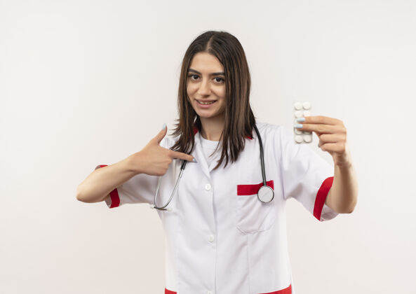 手势年轻的女医生穿着白大褂 脖子上戴着听诊器 站在白色的墙上 用手指指着水泡和药丸人姿势医生