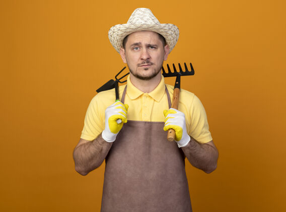 橙色年轻的园丁穿着连体衣 戴着帽子 拿着床垫和小耙子 严肃的脸站在橙色的墙上看着前面工作人床垫