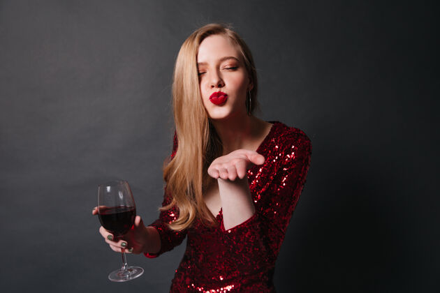 享受戴着酒杯的时髦女士送上了空气之吻摄影棚拍摄的穿着红裙子的金发女孩在派对上喝葡萄酒酒精肖像明亮