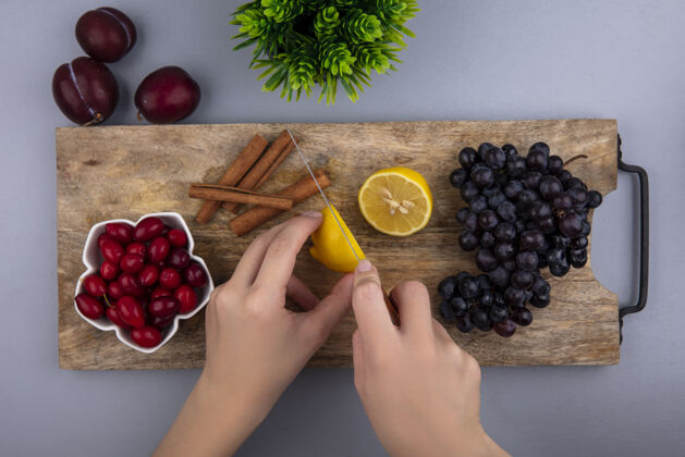 刀俯视图中的女性手切柠檬与刀山茱萸浆果和葡萄肉桂在砧板和普拉特植物的灰色背景雌性柑橘板