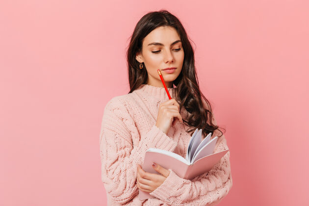 学生穿着粉色毛衣的聪明女人在想她的书的续集深色头发的女人在粉色背景下看她的日记日记头发蓝眼睛