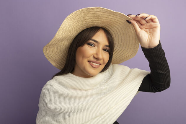 积极戴着白围巾和夏帽的年轻女子站在紫色的墙上 愉快而积极地抚摸着她的帽子围巾紫色女人