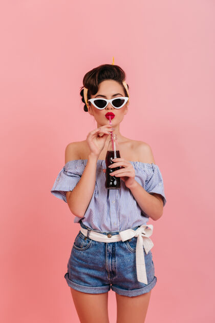 瓶子穿着牛仔短裤和条纹衬衫喝饮料的身材匀称的女孩戴太阳镜的女士的摄影棚照片肖像孤立的粉红色深色