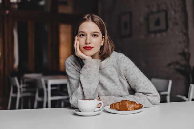 手机咖啡馆里 一个棕色眼睛 穿着羊毛衫的女孩倚在白色的桌子上 看着相机一张红唇女人点咖啡和羊角面包的照片女孩设备人