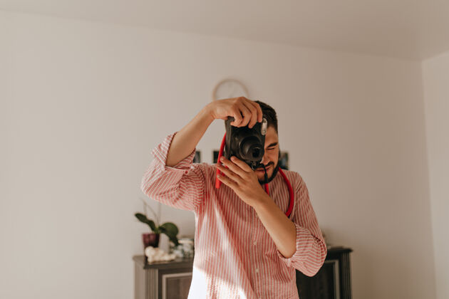 年轻一个手指上戴着戒指的男人在照相机上拍照在宽敞的公寓里 一个身穿条纹服装的深色男人的快照专业时尚衬衫
