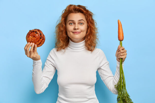 面包房美丽的红发女人在新鲜胡萝卜和甜面包之间选择维生素满意烘焙