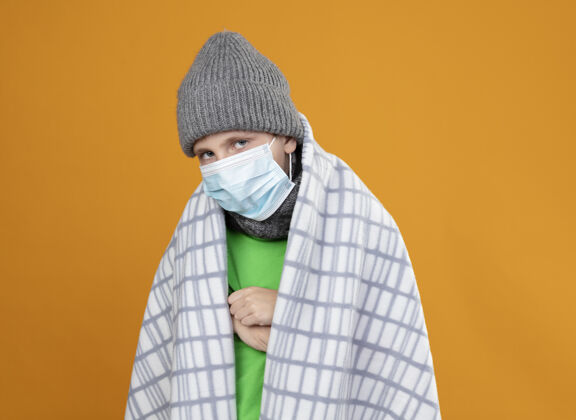 疾病生病的小男孩戴着防护面罩 戴着暖和的帽子 裹着毯子 忍受着翻越橘黄色墙壁的发烧保护包裹痛苦