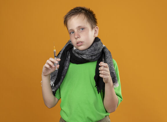 站着生病的小男孩穿着绿色t恤和温暖的围巾感觉不舒服拿着安瓿和注射器站在橙色的墙上不高兴和生病拿着看暖和