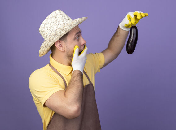 惊人年轻的园丁穿着连体衣 戴着帽子 戴着工作手套 手里拿着茄子 站在紫色的墙上惊讶地看着它帽子人茄子