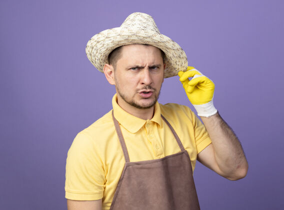 男人年轻的园丁穿着连体衣 戴着工作手套 带着严肃自信的表情站在紫色的墙上看着前方手套工作男性