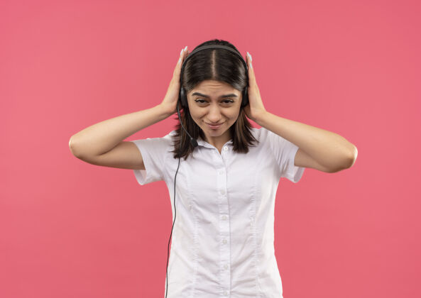 人穿着白衬衫戴着耳机的年轻女孩站在粉红色的墙上 双手合着耳朵 脸上带着恼怒的表情公民麦克风耳朵