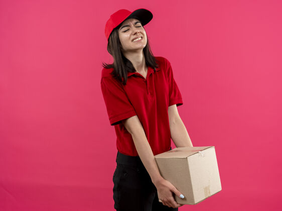马球年轻的送货女孩穿着红色马球衫 戴着帽子 手里拿着沉重的盒装 站在粉色的墙上显得疲惫和紧张盒子包装帽子
