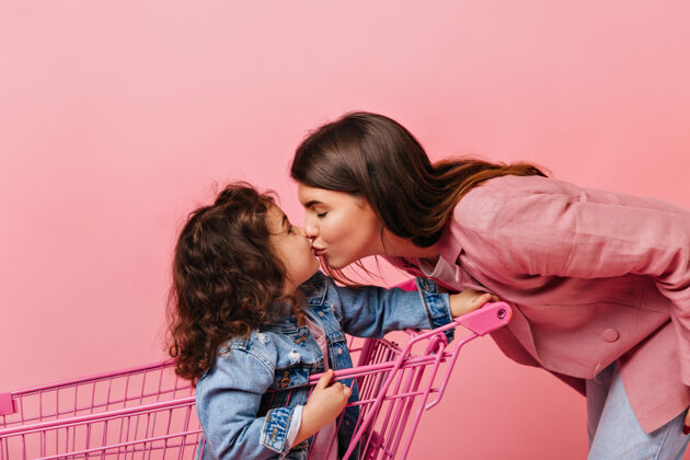女性高兴的年轻女子亲吻卷发的女儿可爱的孩子坐在购物车里表情粉色背景女儿