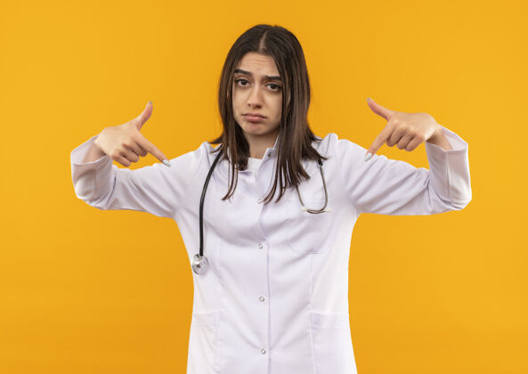 目录年轻的女医生穿着白大褂 脖子上戴着听诊器 用食指指着自己 站在橘色的墙上 脸上带着悲伤的表情 看上去很困惑保健办公室外套