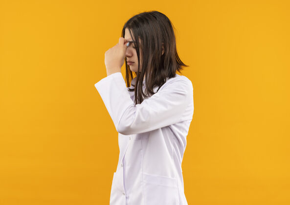 疲倦年轻的女医生穿着白大褂 脖子上戴着听诊器 站在橘黄色的墙上 看上去很疲倦 而且工作过度女性周围办公室