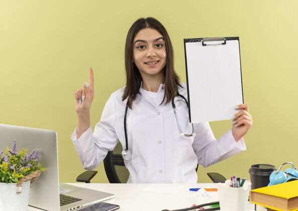 工作身穿白大褂的年轻女医生 脖子上戴着听诊器 手里拿着剪贴板 食指自信地坐在桌旁 笔记本电脑放在浅色的墙上手指拿着剪贴板