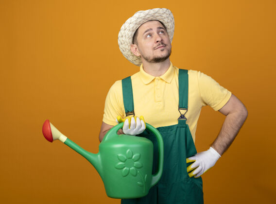 人年轻的园丁穿着连体衣 戴着帽子 站在橘色的墙上困惑地抬头看浇水连身衣人