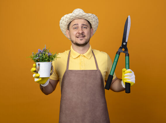 人年轻的园丁穿着连体衣 戴着帽子 拿着树篱剪和盆栽植物 站在橙色的墙上 面带微笑地看着前方植物快乐站着