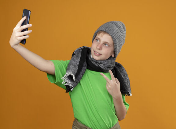 穿生病的小男孩穿着绿色t恤 戴着暖和的围巾和帽子 感觉好多了 拿着智能手机自拍 橙色的墙上有个v型标志男孩Vsign橙色