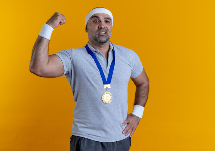 看戴着头巾 脖子上戴着金牌的成熟运动型男人站在橙色的墙上举起拳头 看上去很自信公民男子奖牌