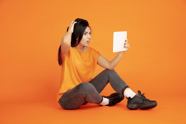 表达橙色工作室背景上的白种人年轻女子肖像穿着衬衫的漂亮黑发女性模特人类情感的概念 面部表情 销售 广告文字空间使用平板电脑 虚拟博客英俊成功年轻
