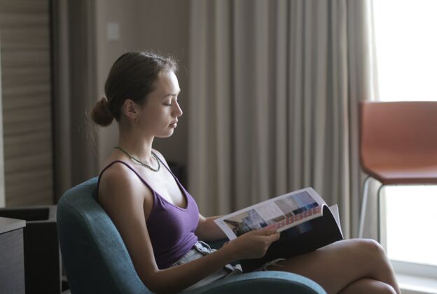 脸一个年轻的女人坐在扶手椅上 在屋里的灯光下看杂志女人休闲年轻人