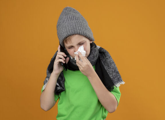 男孩生病的小男孩穿着绿色t恤 戴着暖和的围巾和帽子 不舒服地用手机说话 用纸巾擦鼻子站在橙色的墙上手机温暖穿